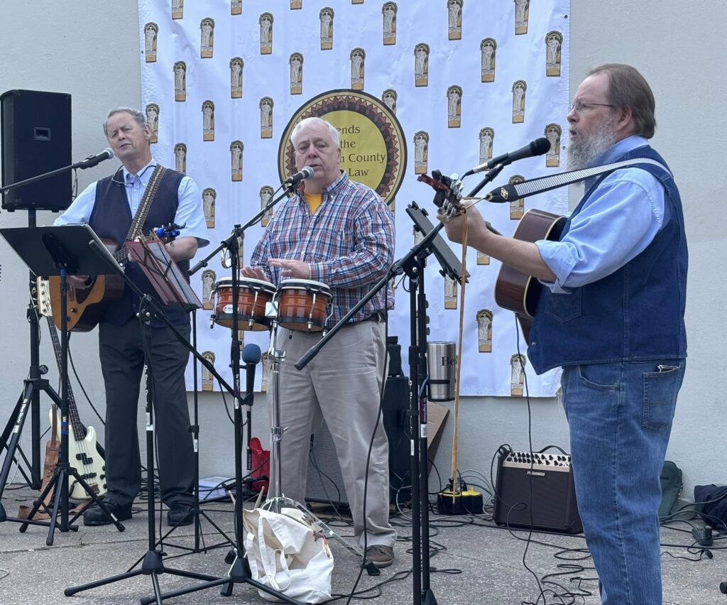 Three older men perform music outdoors; one plays bongos and sings, another plays guitar and sings, and the third plays guitar. They stand in front of a backdrop with a circular logo and a pattern of torch symbols.