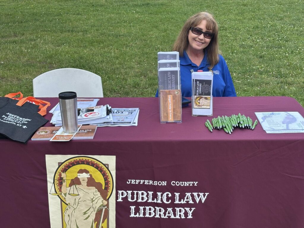 A woman with long brown hair and sunglasses sits at a table covered with a maroon Jefferson County Public Law Library cloth, featuring brochures, pens, tote bags, and other informational materials, outdoors on grass.